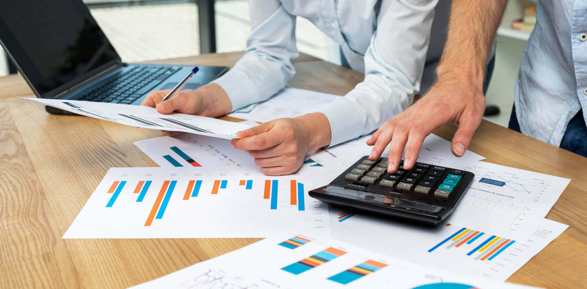 Two persons working on a desk with a calculator, printed charts & laptop placed on it